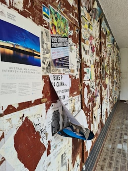 Multiple layers of old and new posters cover a bulletin board in what appears to be a public space. The posters are overlapping and partially torn, giving a cluttered and textured appearance. The floor beside the board is tiled, and there is a rough, textured ceiling.