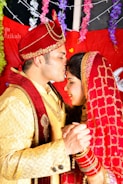 A couple dressed in traditional Indian wedding attire are sharing a tender moment. The man is wearing an ornate red turban and a golden sherwani with red detailing, while the woman is in a red bridal saree with gold embroidery. They are holding hands, and the woman has her eyes closed with a serene expression. The background is decorated with colorful flowers and drapes.