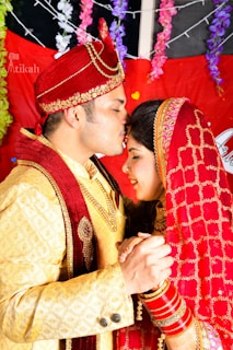 A couple dressed in traditional Indian wedding attire are sharing a tender moment. The man is wearing an ornate red turban and a golden sherwani with red detailing, while the woman is in a red bridal saree with gold embroidery. They are holding hands, and the woman has her eyes closed with a serene expression. The background is decorated with colorful flowers and drapes.