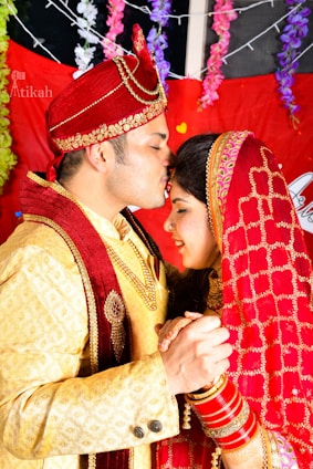 A joyful couple dressed in vibrant Indian wedding attire sharing a tender moment under colorful floral decorations.