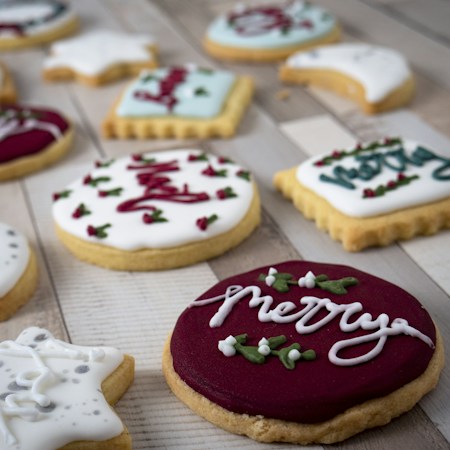 An assortment of decorated sugar cookies, featuring various shapes and icing designs. Some cookies are round with dark red and white icing, others are star or rectangular shaped with light blue icing. The cookies are arranged on a wooden surface, displaying festive messages and decorative patterns like holly leaves.