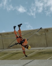 A person wearing an orange safety uniform and helmet is suspended upside down by ropes while performing maintenance work on a building. The sky is clear with scattered clouds.