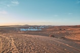 A vast desert landscape with sand dunes extends into the distance, featuring a series of white tents grouped together. The sky shows a gradient from light blue to a warm orange hue near the horizon. Vehicle tire tracks are visible on the sand, suggesting recent human activity.