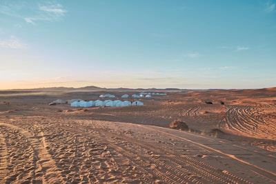 A vast desert landscape with sand dunes extends into the distance, featuring a series of white tents grouped together. The sky shows a gradient from light blue to a warm orange hue near the horizon. Vehicle tire tracks are visible on the sand, suggesting recent human activity.