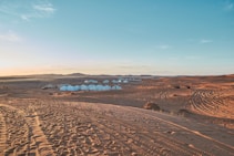A vast desert landscape with sand dunes extends into the distance, featuring a series of white tents grouped together. The sky shows a gradient from light blue to a warm orange hue near the horizon. Vehicle tire tracks are visible on the sand, suggesting recent human activity.