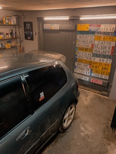 A garage interior featuring a car parked inside. The walls are decorated with a variety of international license plates. A shelf full of tools and car maintenance items is visible on the side. A poster of a well-known fictional character is also on display.