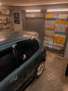A garage interior featuring a car parked inside. The walls are decorated with a variety of international license plates. A shelf full of tools and car maintenance items is visible on the side. A poster of a well-known fictional character is also on display.