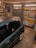 A garage interior featuring a car parked inside. The walls are decorated with a variety of international license plates. A shelf full of tools and car maintenance items is visible on the side. A poster of a well-known fictional character is also on display.