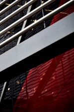 An abstract view of architecture with a focus on geometric shapes and lines. Metal beams run diagonally across the image, with reflections visible on a glossy surface. The background features bold red and black panels with a modern, industrial look.