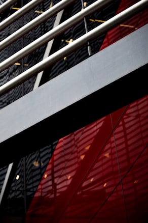 An abstract view of architecture with a focus on geometric shapes and lines. Metal beams run diagonally across the image, with reflections visible on a glossy surface. The background features bold red and black panels with a modern, industrial look.