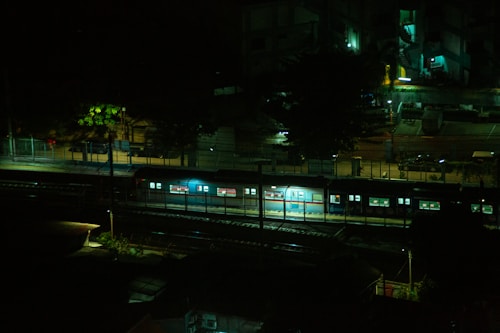 A night scene featuring a train stopped on tracks, with dim lighting illuminating parts of the train and surrounding area. Buildings and streetlights are visible in the background, casting a mix of shadows and glowing lights.