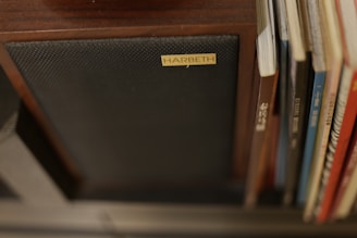 A speaker with a label reading 'Harbeth' is positioned next to a row of books or vinyl records. The wooden finish of the speaker and dark-colored fabric cover can be seen, alongside the spines of various books or records in assorted colors and styles.