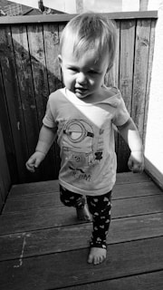 A young child walking on a wooden deck, wearing a t-shirt with a cartoon character and pants with heart patterns. The child is barefoot and the image is in black and white.
