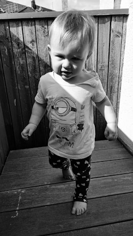 A young child walking on a wooden deck, wearing a t-shirt with a cartoon character and pants with heart patterns. The child is barefoot and the image is in black and white.