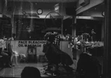 A monochrome image of a barbershop interior. The shop is busy with people, and a barber appears to be cutting a customer's hair. The reflection in the mirror shows multiple chairs lined with various haircare products. A sign on the window advertises services like face bleaching and oil massage.