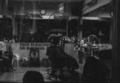 A monochrome image of a barbershop interior. The shop is busy with people, and a barber appears to be cutting a customer's hair. The reflection in the mirror shows multiple chairs lined with various haircare products. A sign on the window advertises services like face bleaching and oil massage.