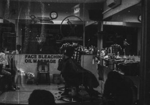 A monochrome image of a barbershop interior. The shop is busy with people, and a barber appears to be cutting a customer's hair. The reflection in the mirror shows multiple chairs lined with various haircare products. A sign on the window advertises services like face bleaching and oil massage.