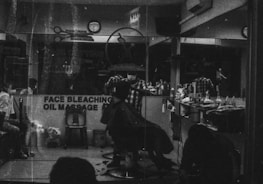 A monochrome image of a barbershop interior. The shop is busy with people, and a barber appears to be cutting a customer's hair. The reflection in the mirror shows multiple chairs lined with various haircare products. A sign on the window advertises services like face bleaching and oil massage.