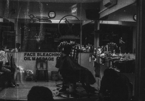 A monochrome image of a barbershop interior. The shop is busy with people, and a barber appears to be cutting a customer's hair. The reflection in the mirror shows multiple chairs lined with various haircare products. A sign on the window advertises services like face bleaching and oil massage.