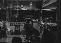 A monochrome image of a barbershop interior. The shop is busy with people, and a barber appears to be cutting a customer's hair. The reflection in the mirror shows multiple chairs lined with various haircare products. A sign on the window advertises services like face bleaching and oil massage.