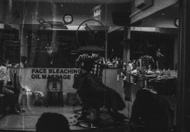 A monochrome image of a barbershop interior. The shop is busy with people, and a barber appears to be cutting a customer's hair. The reflection in the mirror shows multiple chairs lined with various haircare products. A sign on the window advertises services like face bleaching and oil massage.