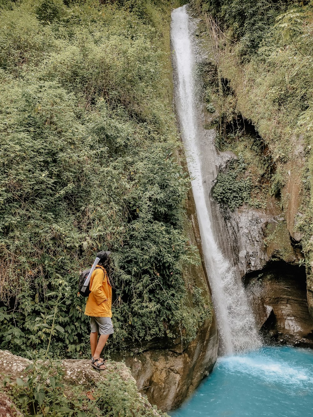 man in orange shirt and blue shorts standing on rock near waterfalls during daytime