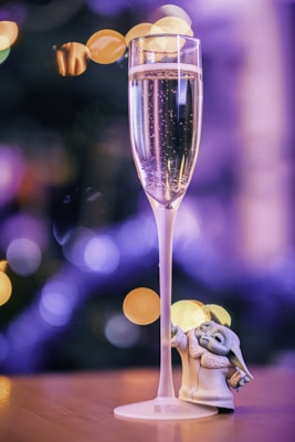 A tall, elegant champagne flute filled with bubbling sparkling wine is prominently featured, with soft bokeh lights in the background providing a festive and dreamy atmosphere. Next to the glass is a small, detailed figurine resembling a fictional character, which adds an element of whimsy to the scene.