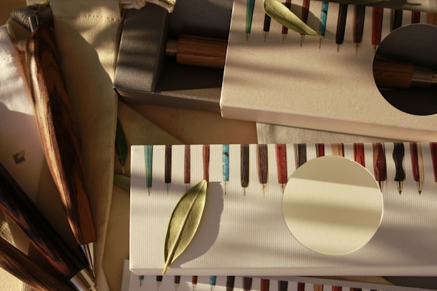 A collection of exquisitely crafted wooden pens displayed in elegant packaging, with a leaf delicately placed on top. The pens have a variety of colorful and glossy patterns, each with a distinct appearance. The soft, warm lighting creates gentle shadows, adding depth and character to the scene.