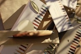 An assortment of writing instruments, including a wooden pen, is arranged on neutral-colored fabric and paper. Small green leaves are scattered among the items, and the scene is bathed in soft, natural light with shadows of branches visible.