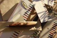 An assortment of writing instruments, including a wooden pen, is arranged on neutral-colored fabric and paper. Small green leaves are scattered among the items, and the scene is bathed in soft, natural light with shadows of branches visible.