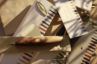 An assortment of writing instruments, including a wooden pen, is arranged on neutral-colored fabric and paper. Small green leaves are scattered among the items, and the scene is bathed in soft, natural light with shadows of branches visible.