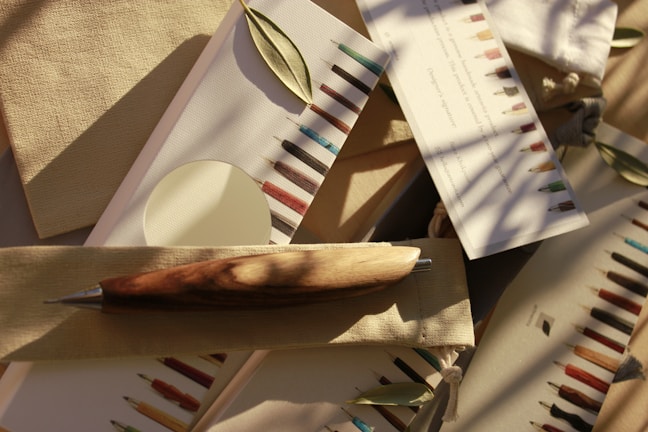 An assortment of writing instruments, including a wooden pen, is arranged on neutral-colored fabric and paper. Small green leaves are scattered among the items, and the scene is bathed in soft, natural light with shadows of branches visible.