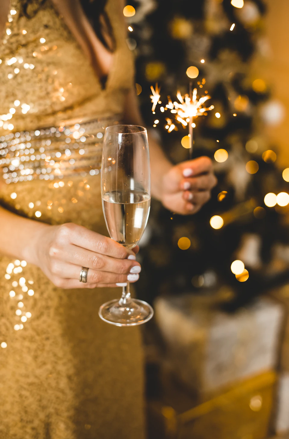 The image shows a person dressed in a gold sequined dress holding a champagne flute filled with champagne, which is being sparkled by a small firecracker. The background features a Christmas tree adorned with gold and white ornaments against a gray wall.