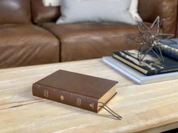 A wooden coffee table with a brown book titled 'Holy Bible' placed on it. The cover of the Bible is leather. Next to it, there are stacked books and a geometric decorative object. In the background, a brown leather couch with a pillow is partially visible.