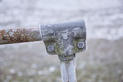 A close-up view of a metal pipe joint, covered with a thin layer of frost. The metal appears weathered with rust and some green moss growth. The background is blurred, giving prominence to the frosty texture on the pipe.