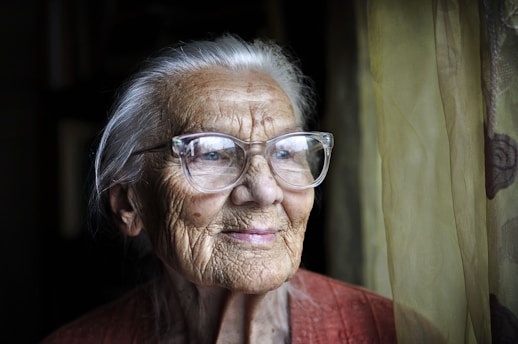 An elderly person with silver hair and large glasses gazes thoughtfully out a window. The ambient lighting softly highlights the person's facial wrinkles and expression. A muted curtain forms the backdrop, creating a peaceful and reflective atmosphere.