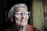 An elderly person with silver hair and large glasses gazes thoughtfully out a window. The ambient lighting softly highlights the person's facial wrinkles and expression. A muted curtain forms the backdrop, creating a peaceful and reflective atmosphere.