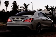 A sleek, white Mercedes-Benz AMG car is parked on a street with palm trees in the background. The car's rear lights are illuminated, indicating it's either starting or stopping. The setting appears to be at dusk with the sky darkening, enhancing the contrast of the car's lights.