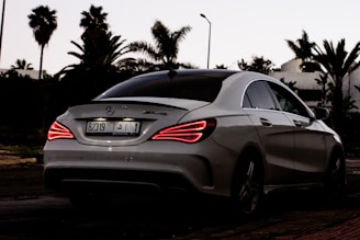 A sleek, white Mercedes-Benz AMG car is parked on a street with palm trees in the background. The car's rear lights are illuminated, indicating it's either starting or stopping. The setting appears to be at dusk with the sky darkening, enhancing the contrast of the car's lights.
