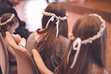 Several young girls with long hair are seated and wearing floral headbands with ribbons. The atmosphere suggests a formal event or ceremony. The background is softly blurred, creating a serene and gentle ambiance.