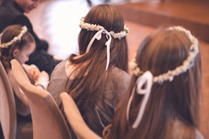 Several young girls with long hair are seated and wearing floral headbands with ribbons. The atmosphere suggests a formal event or ceremony. The background is softly blurred, creating a serene and gentle ambiance.