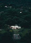 Aerial view of a house surrounded by dense green forest. The house features a flat roof, multiple levels, and a mix of white and red colors. In front of the house, there is a tennis court with a worn surface. The area is lush with various trees creating a secluded and serene atmosphere.