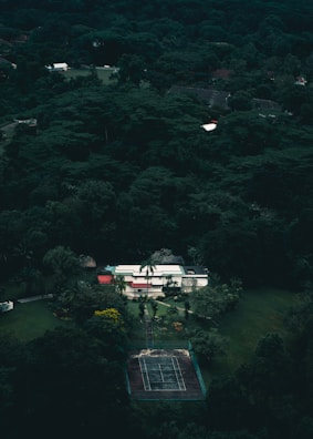 Aerial view of a house surrounded by dense green forest. The house features a flat roof, multiple levels, and a mix of white and red colors. In front of the house, there is a tennis court with a worn surface. The area is lush with various trees creating a secluded and serene atmosphere.