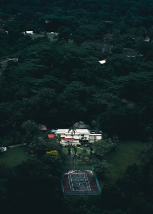 Aerial view of a house surrounded by dense green forest. The house features a flat roof, multiple levels, and a mix of white and red colors. In front of the house, there is a tennis court with a worn surface. The area is lush with various trees creating a secluded and serene atmosphere.