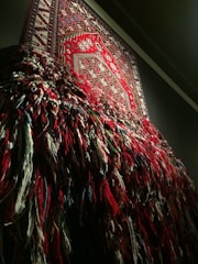 An intricately woven tapestry with a detailed geometric pattern is displayed on a wall. The lower half of the tapestry has loose, multicolored threads hanging down, creating a textured and layered appearance.