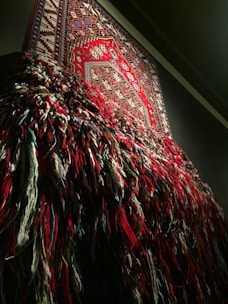 An intricately woven tapestry with a detailed geometric pattern is displayed on a wall. The lower half of the tapestry has loose, multicolored threads hanging down, creating a textured and layered appearance.