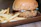 A juicy fried chicken sandwich sits on a wooden tray alongside a serving of golden, crispy french fries. The sandwich is topped with cheese and a light drizzle of a creamy orange sauce, all encased in a soft, toasted bun.