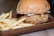 A juicy fried chicken sandwich sits on a wooden tray alongside a serving of golden, crispy french fries. The sandwich is topped with cheese and a light drizzle of a creamy orange sauce, all encased in a soft, toasted bun.