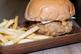A juicy fried chicken sandwich sits on a wooden tray alongside a serving of golden, crispy french fries. The sandwich is topped with cheese and a light drizzle of a creamy orange sauce, all encased in a soft, toasted bun.