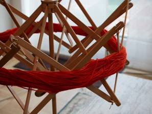 A wooden yarn swift holds a vibrant red skein of yarn, featuring a geometric arrangement of wooden arms that support the yarn in a circular shape. The setup is located indoors, likely in a workspace for knitting or weaving.